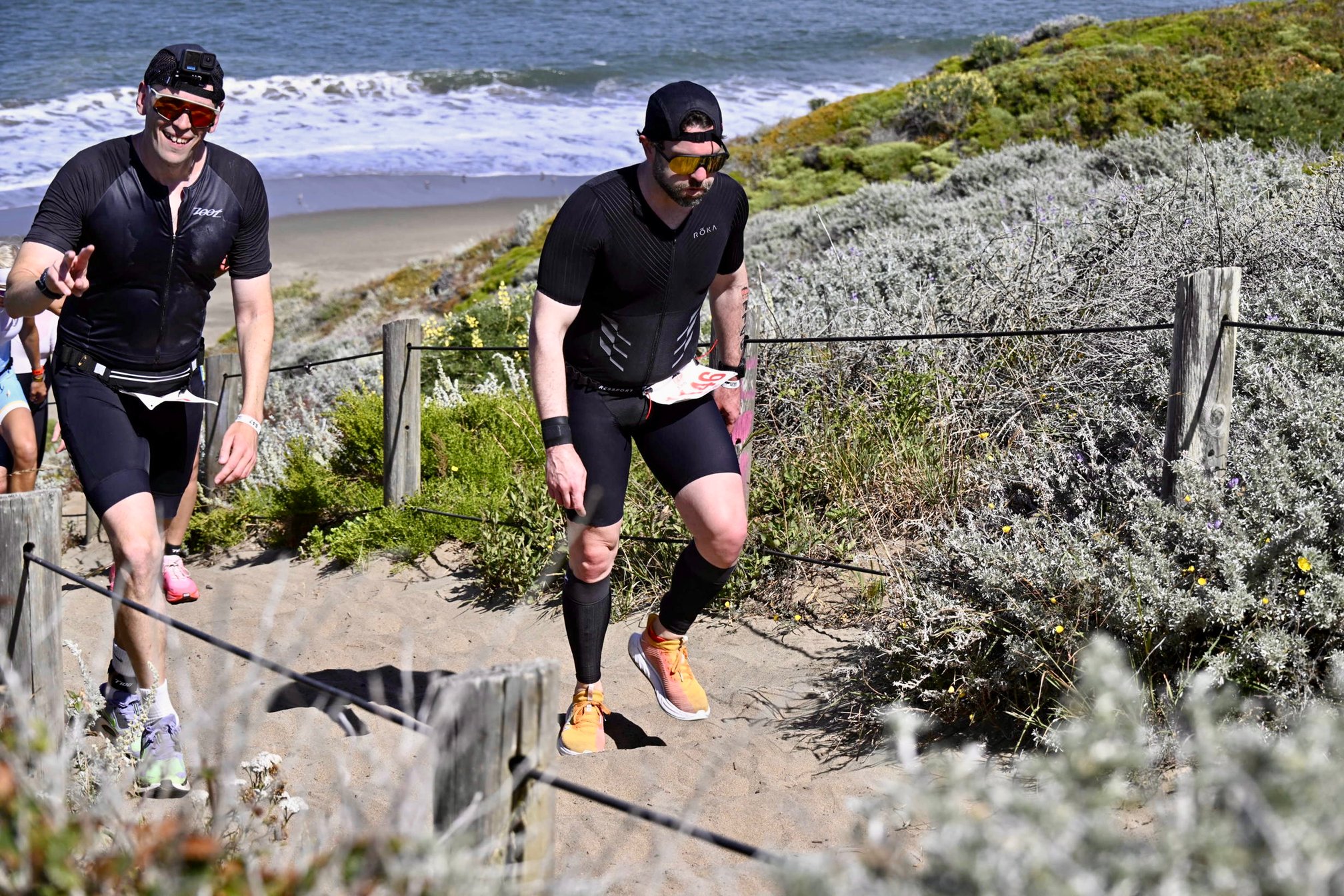 Guillermo Esteves, wearing a black trisuit, backwards hat, calf sleeves, and yellow shoes, climbing the Sand Ladder at Baker Beach, with a look of exhaustion on his face. Behind him, another triathlete wearing a black trisuit with a GoPro strapped to his forehead smiles and flashes a peace sign at the camera.