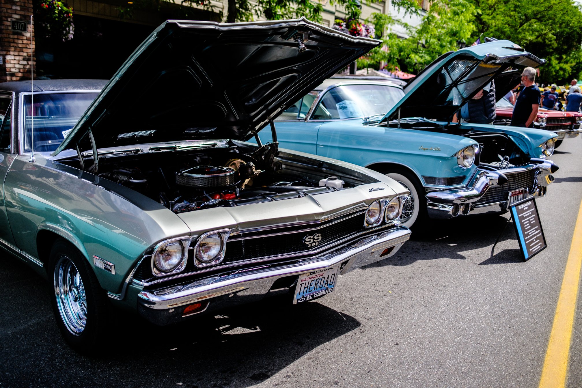 Several classic cars displayed on a street, with the hoods open. In the foreground, a green Chevrolet Chevelle SS 396 with a "THEROAD" Washington state license plate; next to it, a teal Cadillac Coupe de Ville.