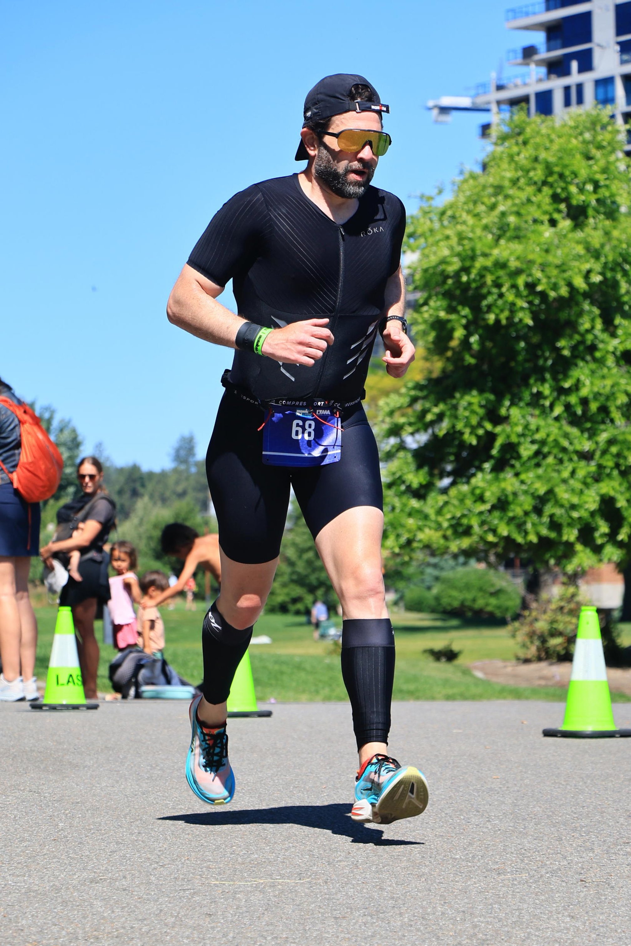 Guillermo Esteves running on a paved path, wearing a black cap, sunglasses, a black trisuit with race bib 68, and calf sleeves.