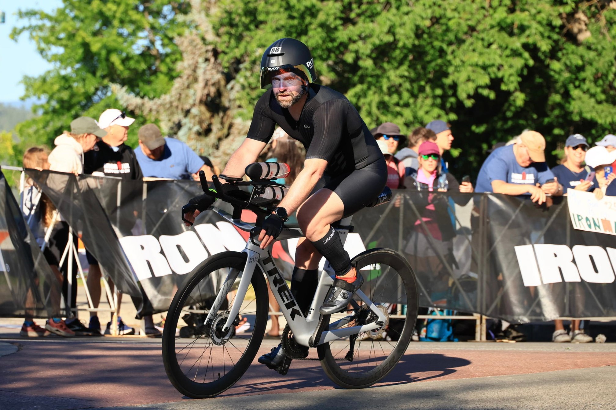 Guillermo Esteves riding a silver Trek Speed Concept bike, wearing a black aero helmet with number 68, a black trisuit, and bike gloves, riding on the base bars. Spectators stand behind Ironman barriers in the background.