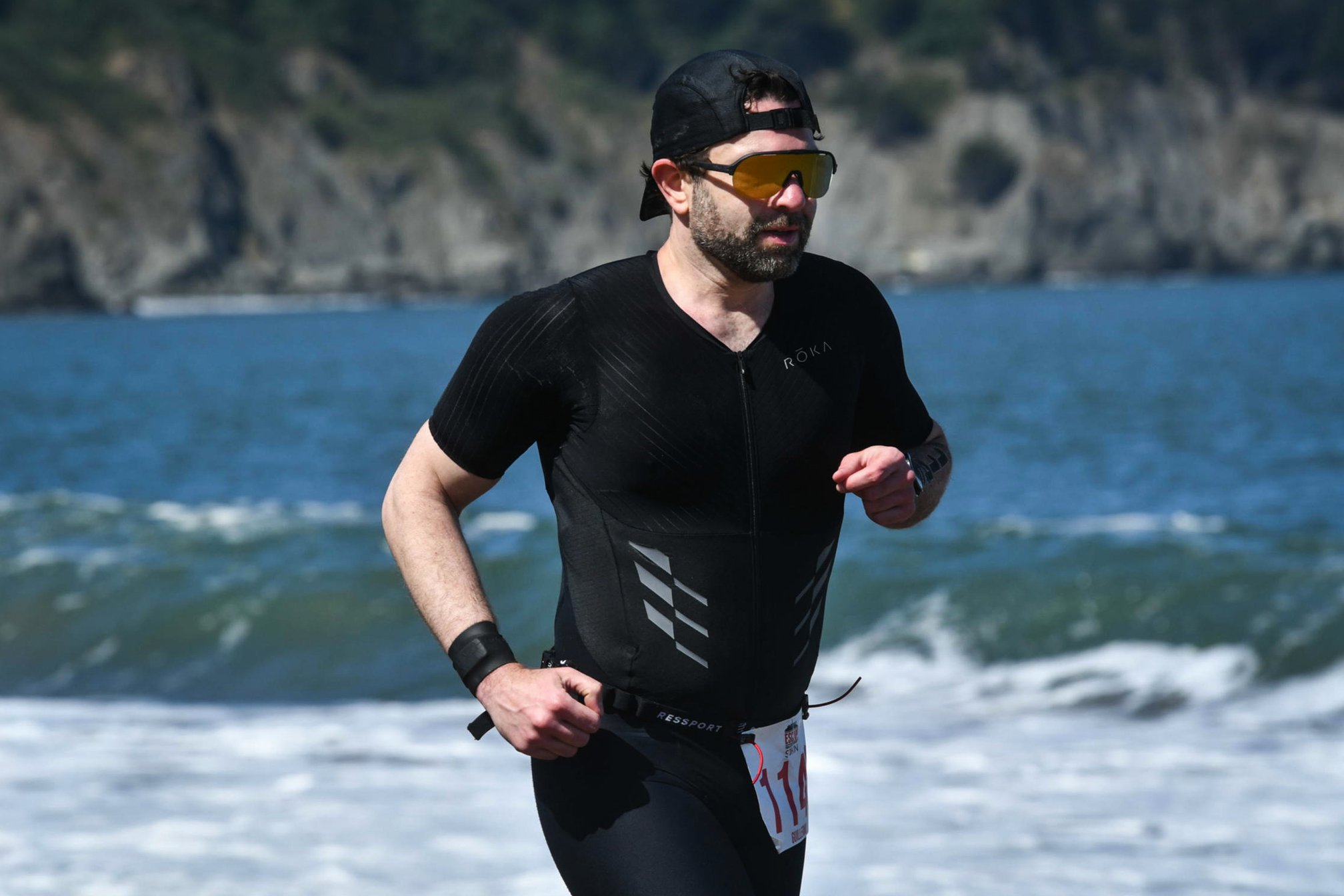 Guillermo Esteves running along Baker Beach in a black trisuit, backward cap, and sunglasses. Waves break behind him below a steep rocky headland.