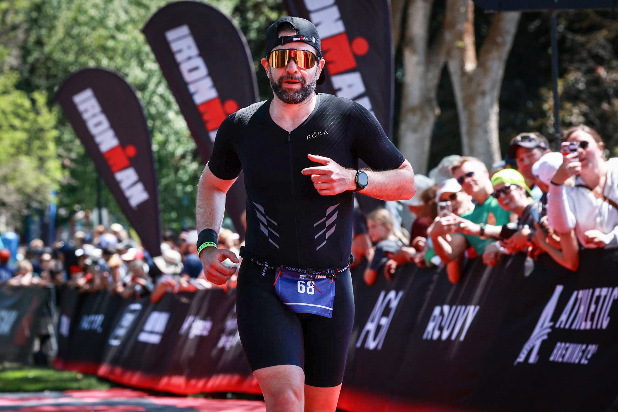 Guillermo Esteves running near the finish line, wearing a black cap, mirrored sunglasses, and a black trisuit with race bib 68. Ironman flags and a crowd of spectators behind a barrier appear in the background.