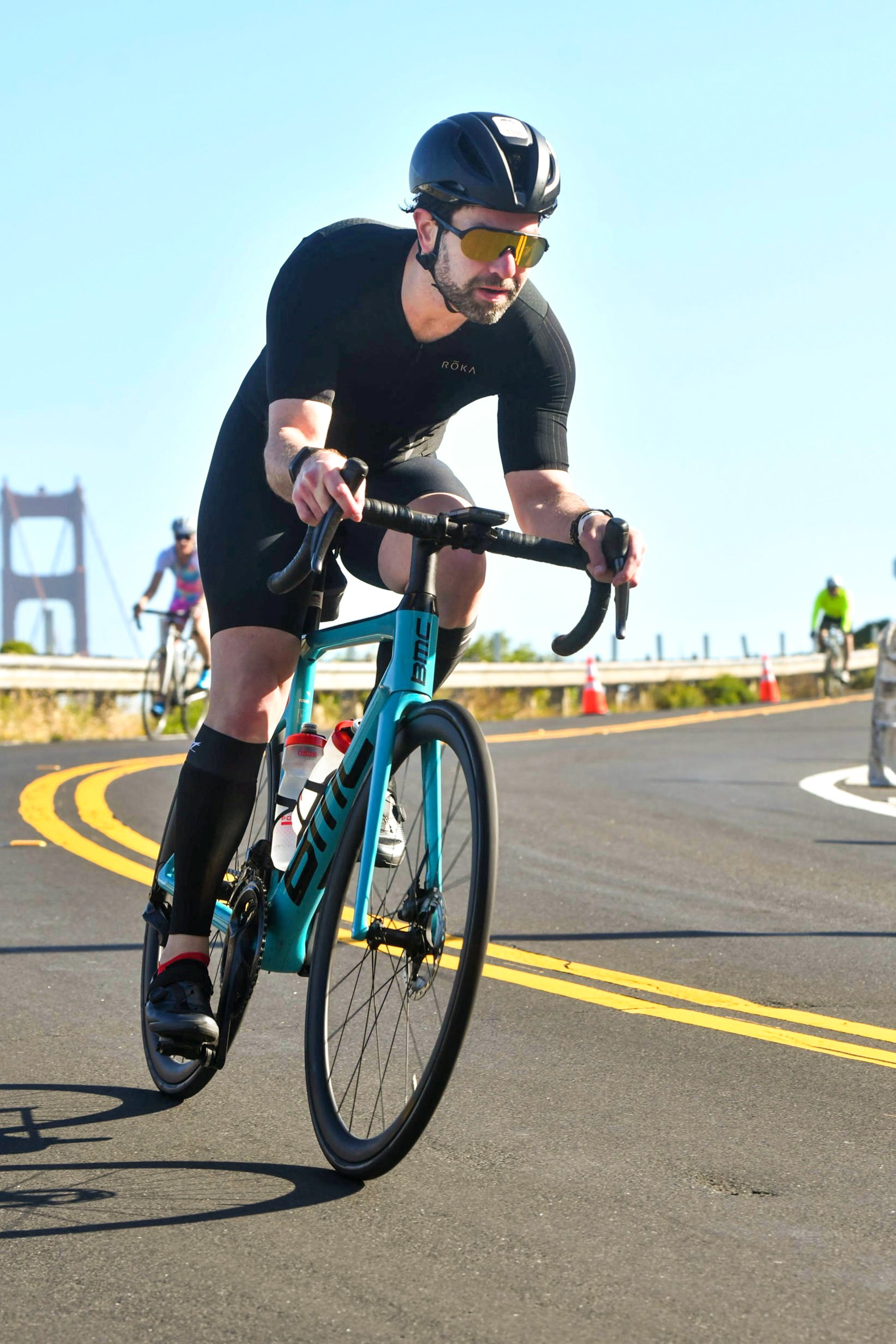 Guillermo Esteves, wearing a black trisuit, helmet, and sunglasses riding a teal BMC road bike toward the camera on a curving road, with a tower of the Golden Gate Bridge in the background.