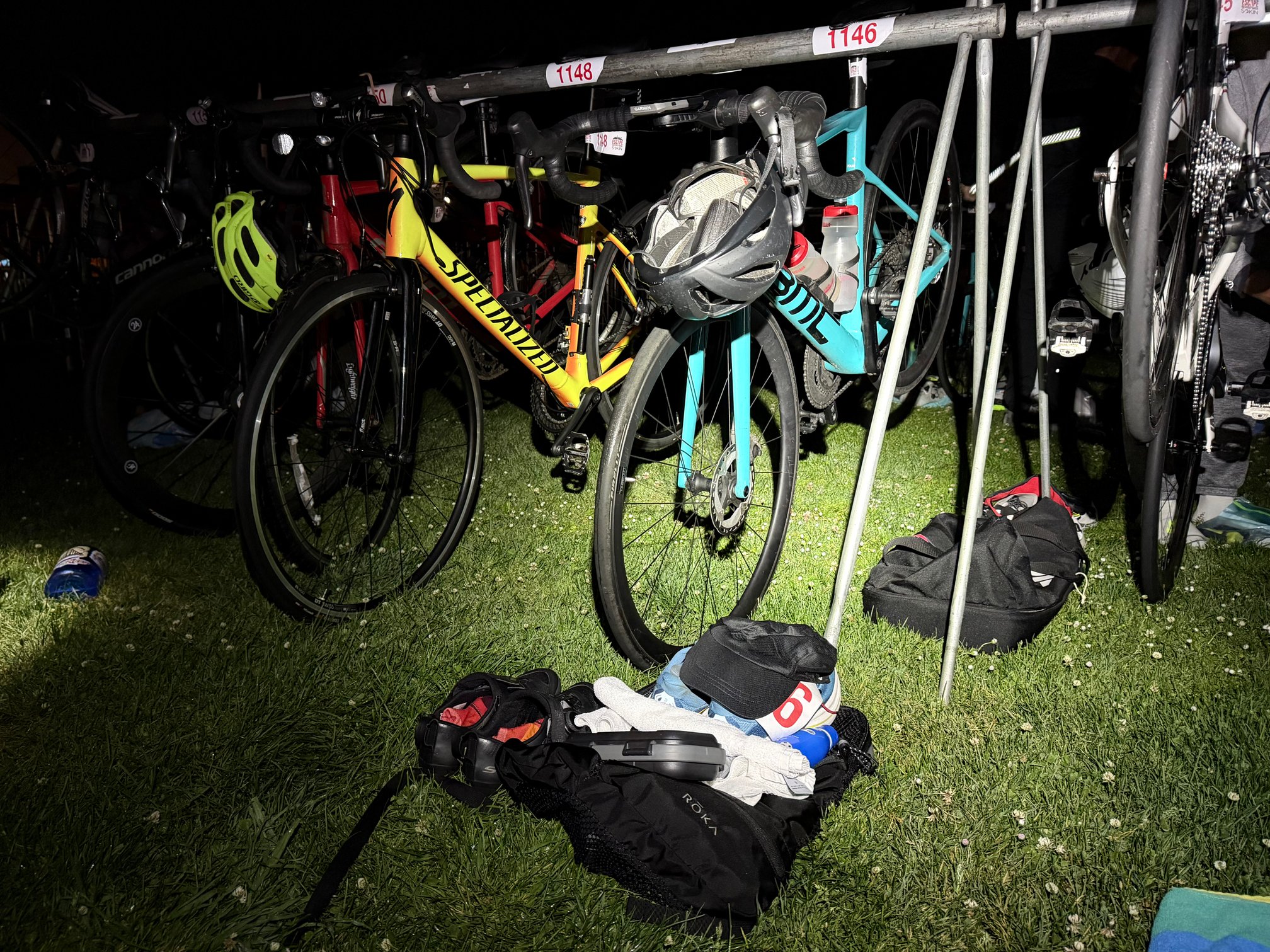 Road bikes hanging by their saddles from a metal transition rack before dawn, with race number stickers reading 1146 and 1148. A yellow Specialized bike and a teal BMC bike are in the foreground, lit by a headlamp, with a helmet hooked over the handlebars. Transition bags, towels, cycling shoes, and other gear are spread out on the grass below.