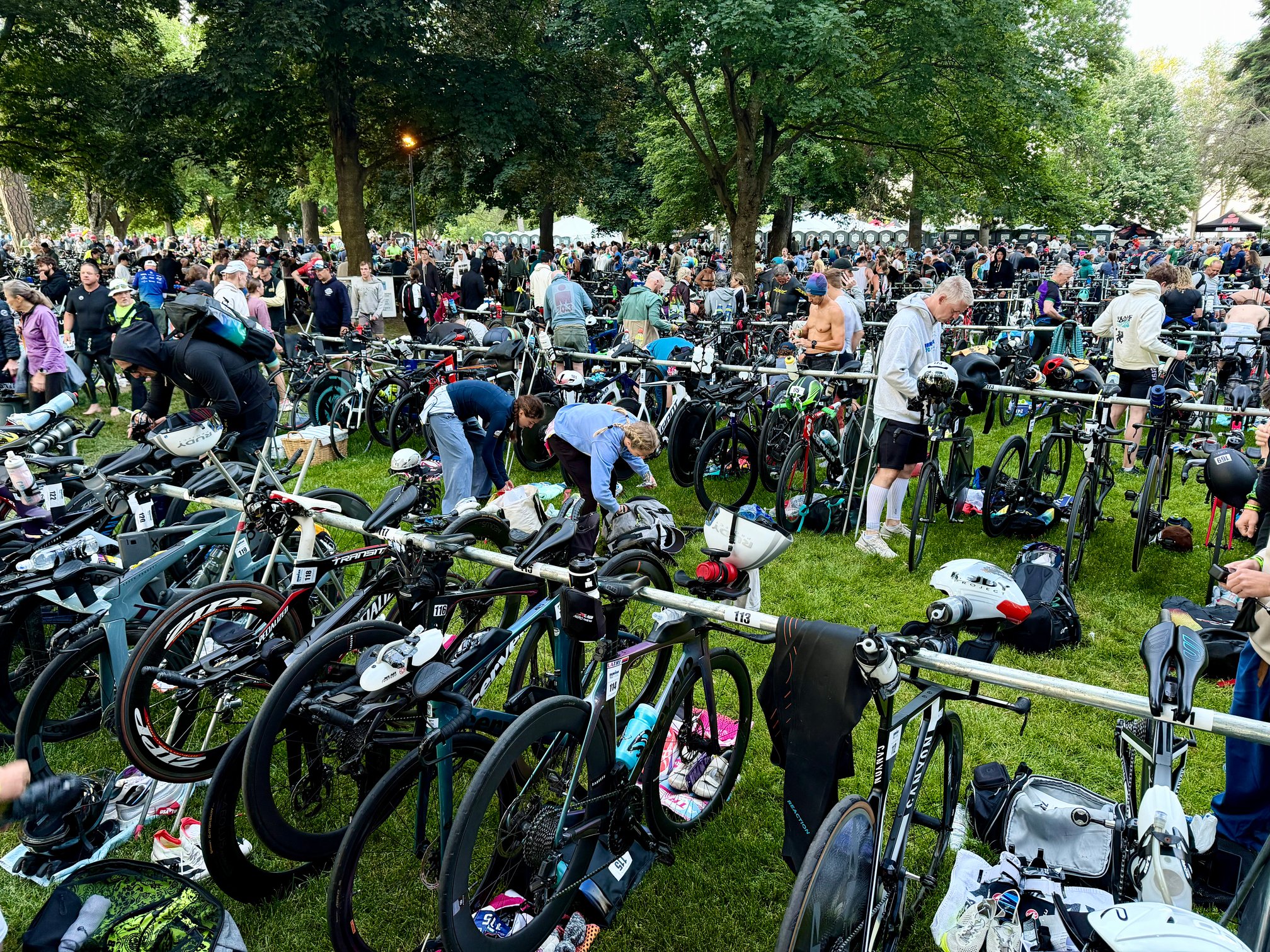 A crowded triathlon transition area in a tree-shaded park, with rows of bikes racked on metal rails. Athletes are setting up their gear, with numbered bib tags visible on the rails.