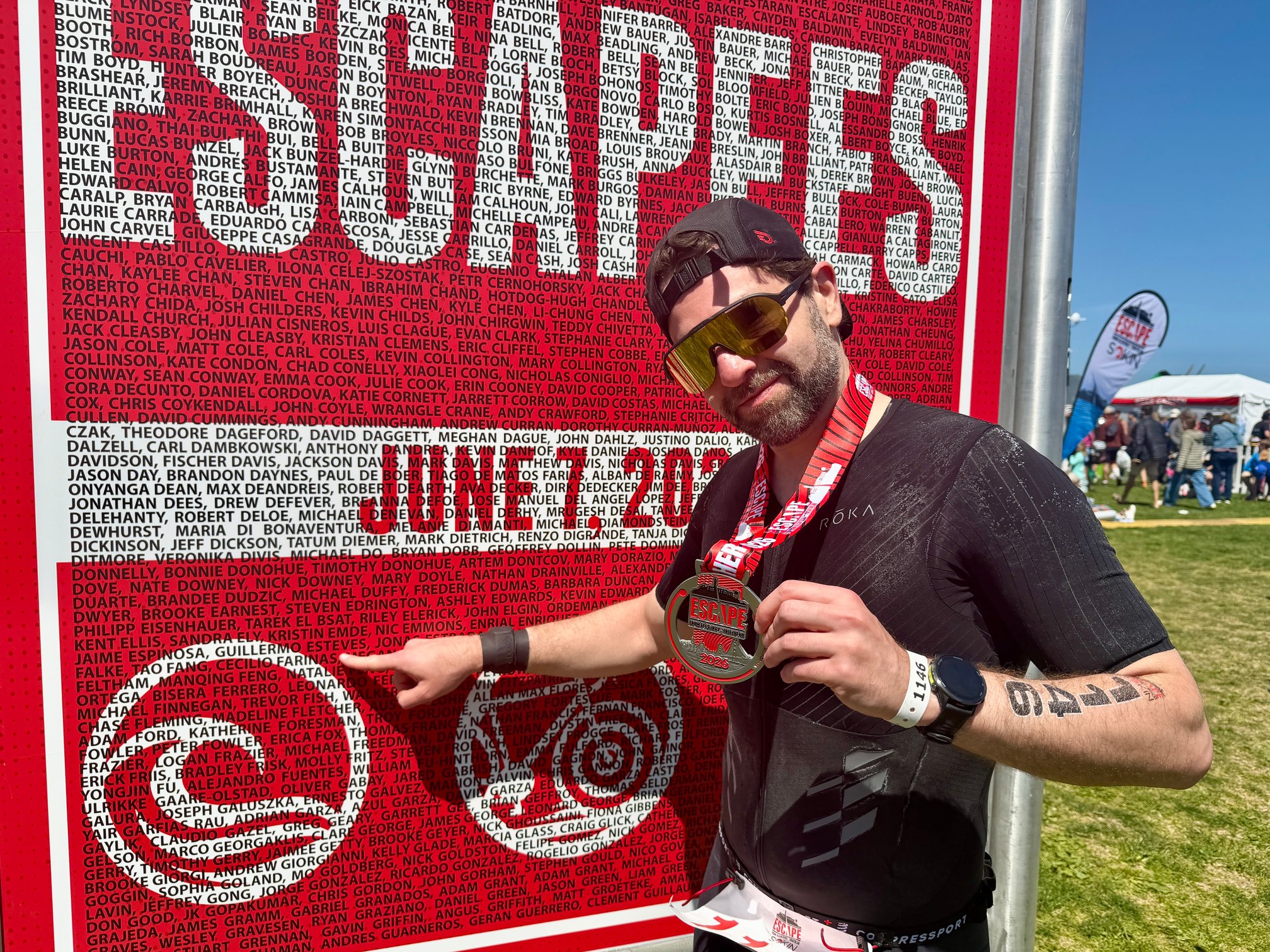 Guillermo Esteves, wearing a black trisuit, backwards cap, and sunglasses, standing in front of a large red finisher board covered in participants' names, holding up an Escape From Alcatraz 2026 finisher medal and pointing at his own name name on the board.