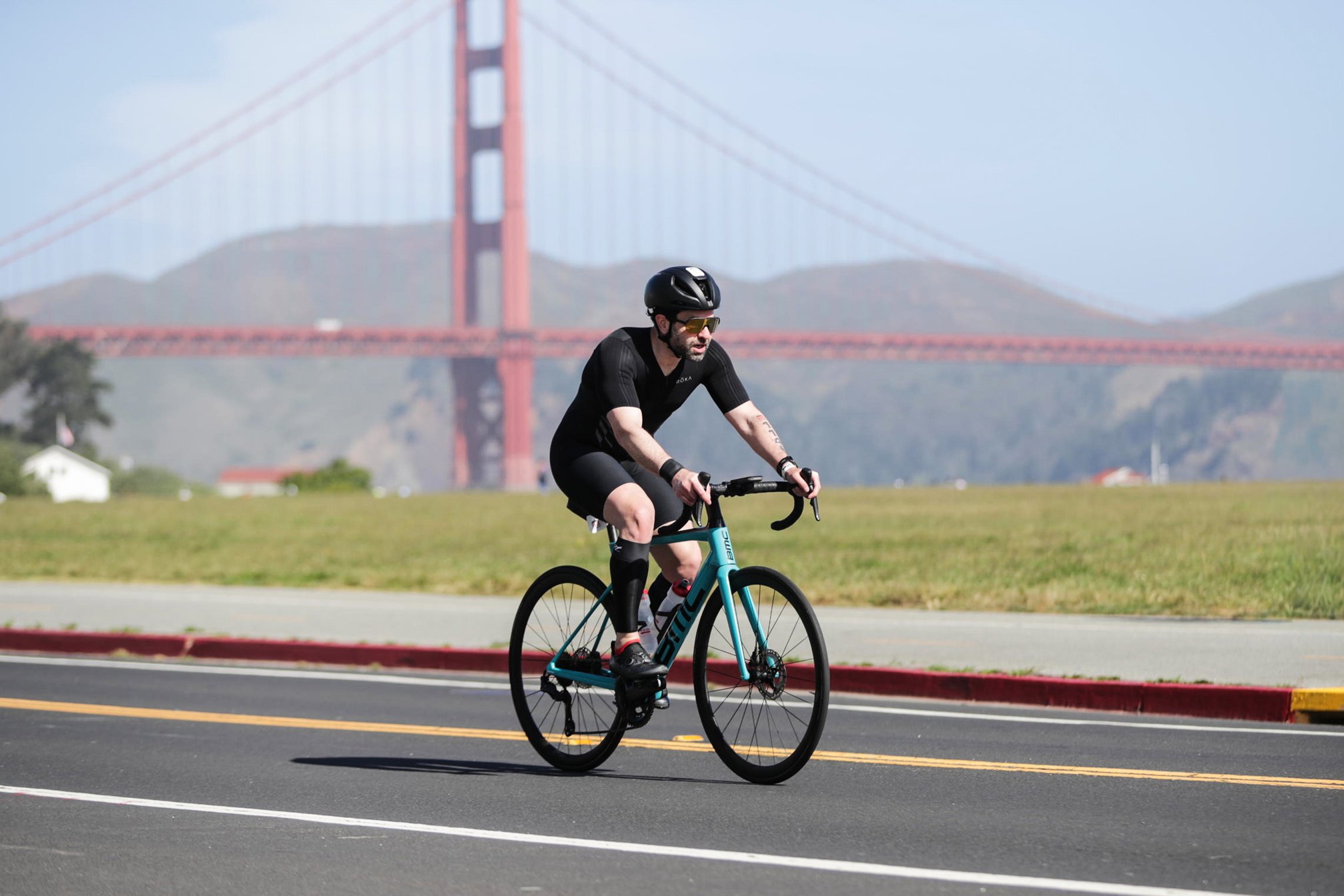 Guillermo Esteves, wearing a black trisuit, helmet, and sunglasses, riding a teal BMC road bike along Crissy Field, with the Golden Gate Bridge and headlands rising in the background.
