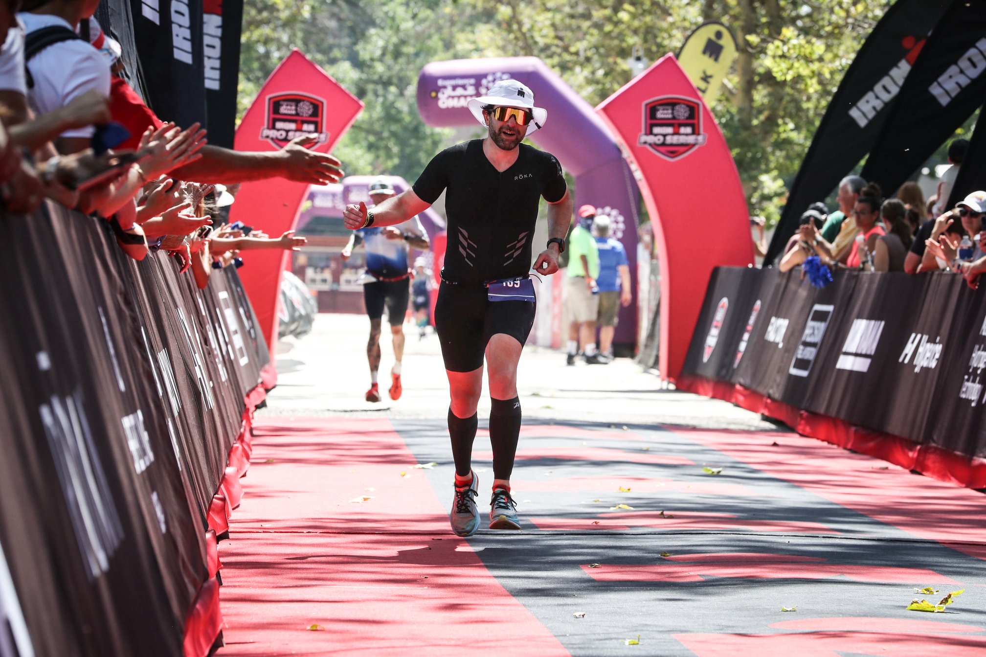 Guillermo Esteves, wearing a white boonie hat, sunglasses, and black trisuit running along the finish chute of Ironman 70.3 Boise, with spectators lining both sides of the red-carpeted course and cheering.