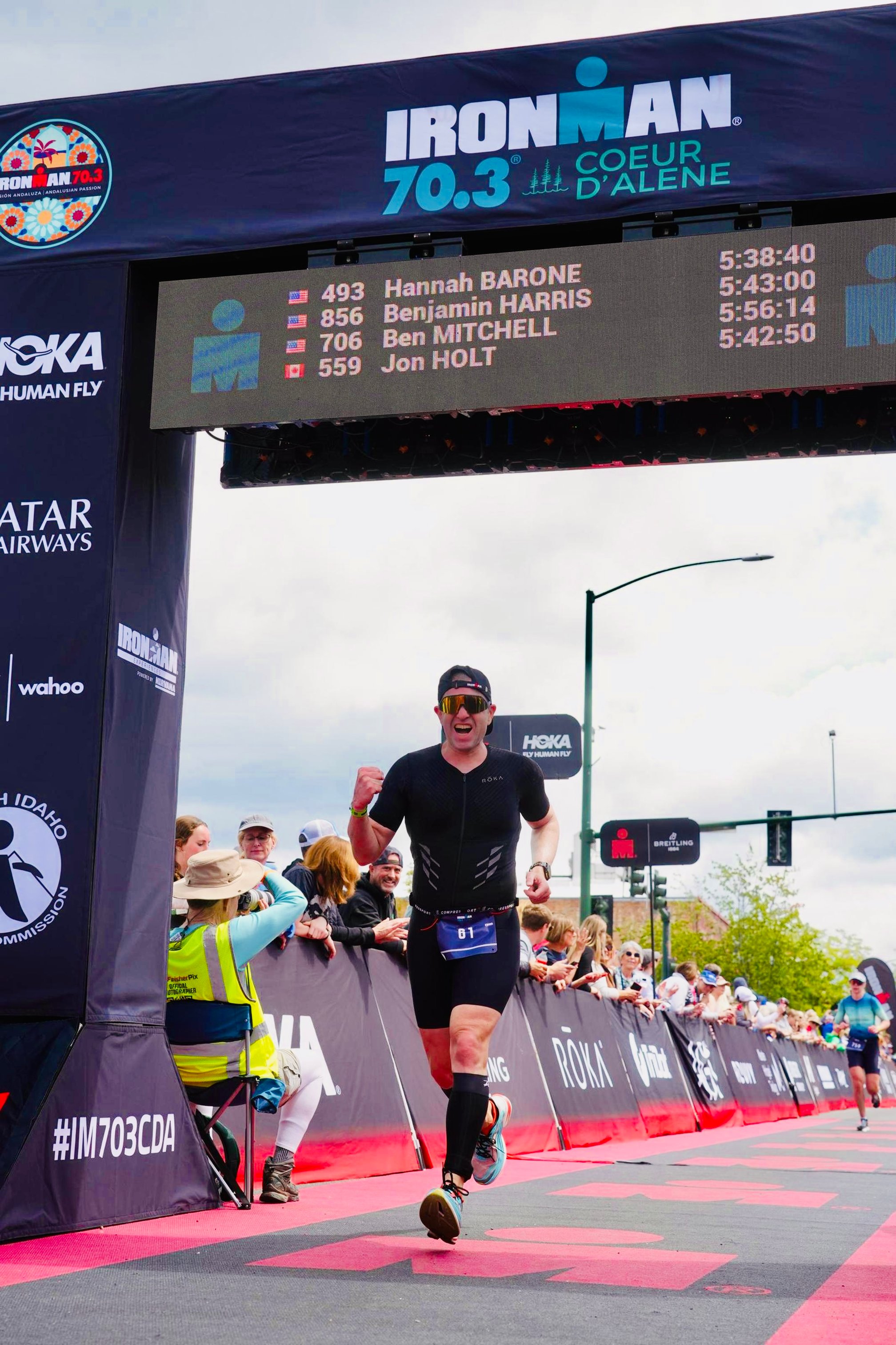 Guillermo Esteves, wearing a race bib number 61, running across the finish line of Ironman 70.3 Coeur d'Alene, with a digital leaderboard above displaying finish times and names.
