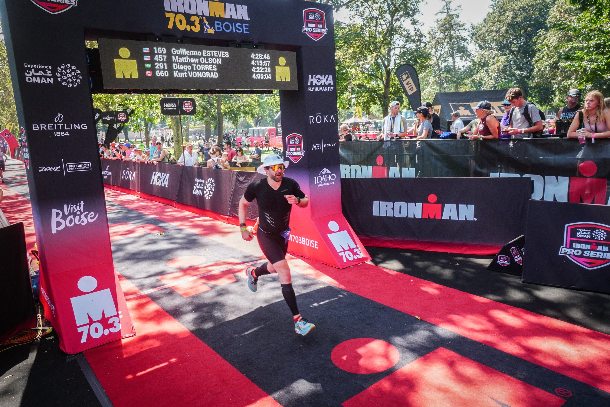 Guillermo Esteves, wearing a black trisuit, calf sleeves, and a white boonie hat, crossing the finish line at Ironman 70.3 Boise, running beneath a leaderboard displaying race times and the Ironman logo, with spectators visible in the background behind barriers.