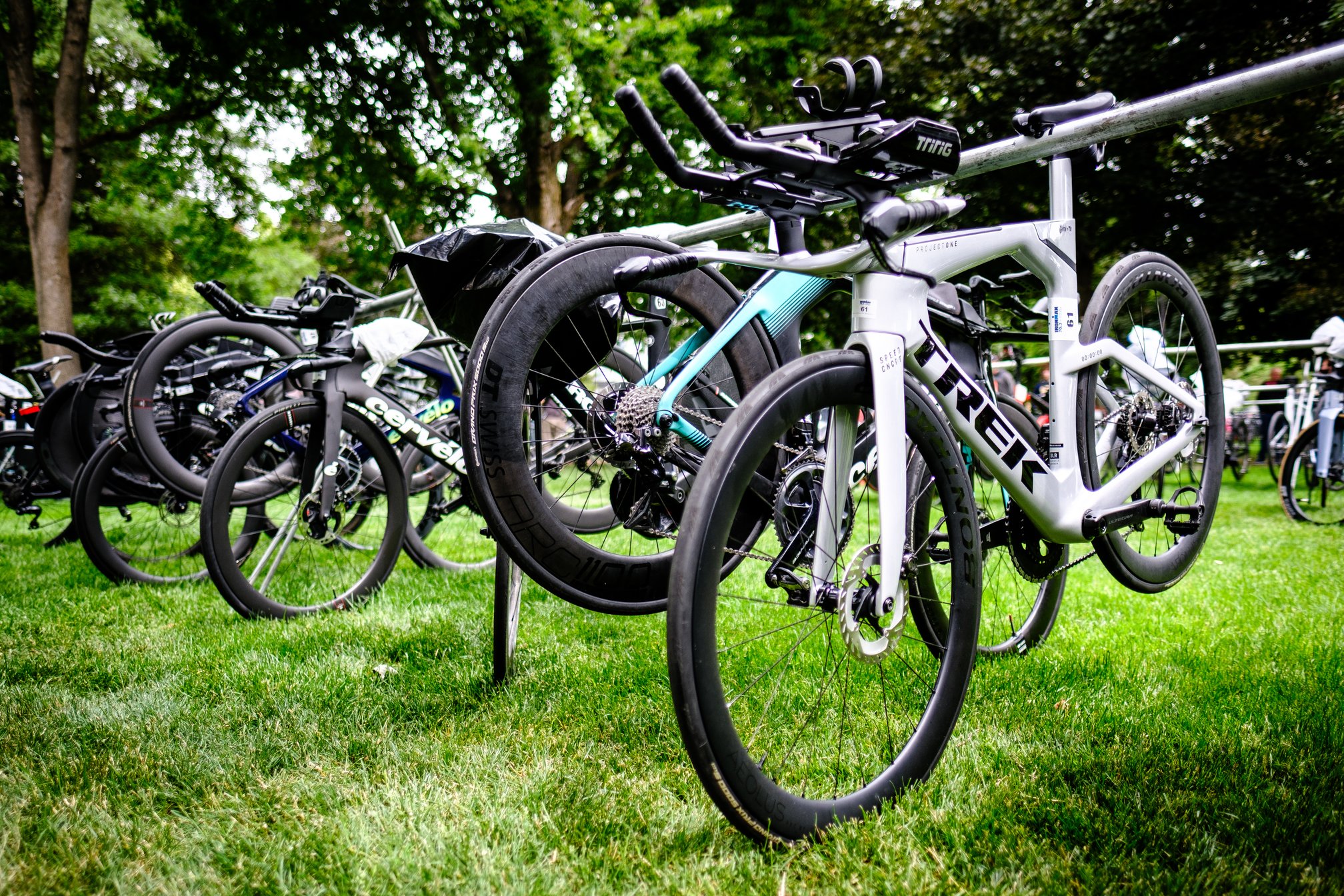 A silver Trek Speed Concept bike racked on a metal bar in a grassy transition area, surrounded by trees and other racked bikes.