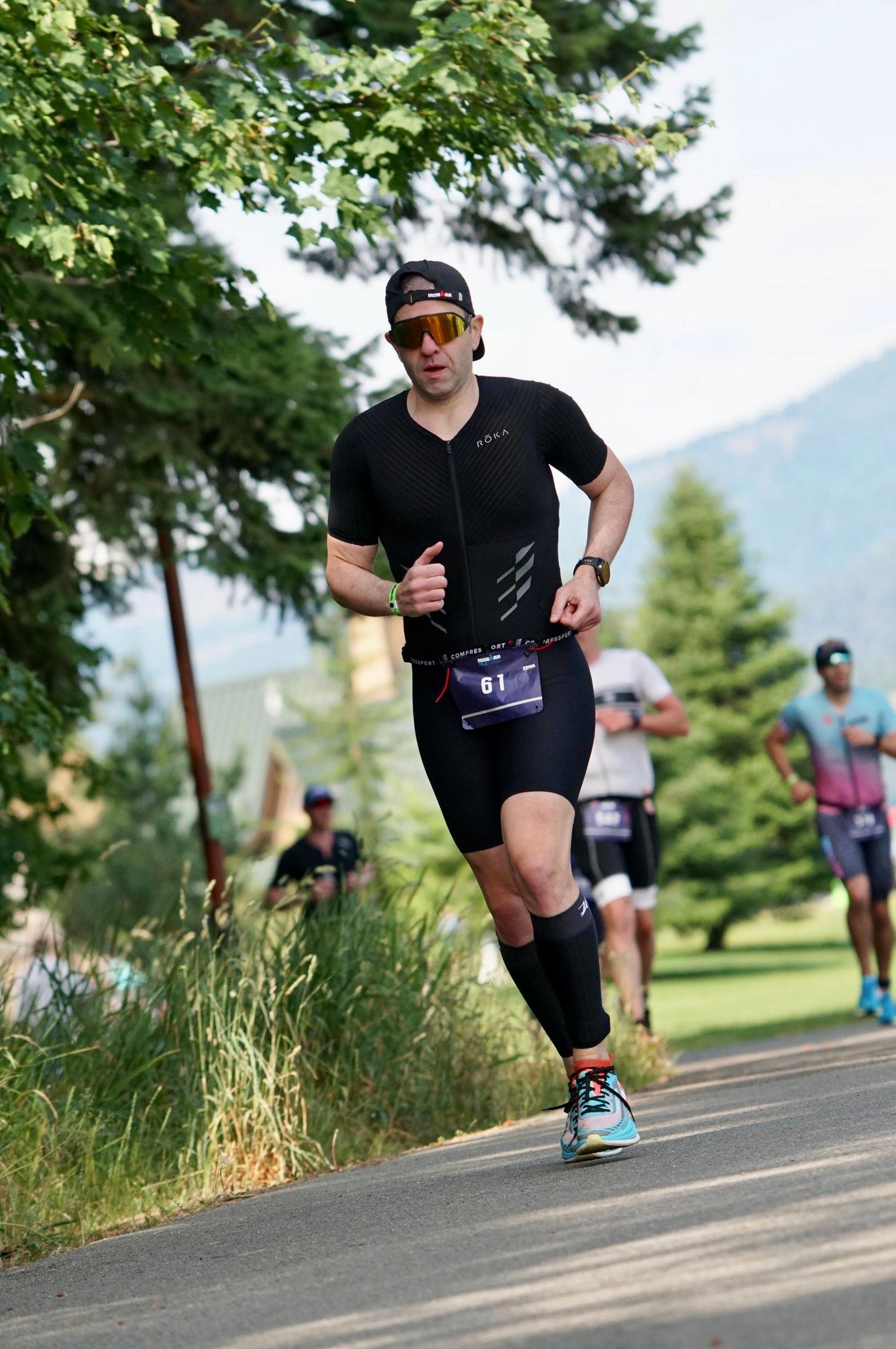 Guillermo Esteves, wearing a black trisuit, race bib number 61, and sunglasses, running on a tree-lined paved path during Ironman 70.3 Coeur d'Alene, with several other runners in the background.