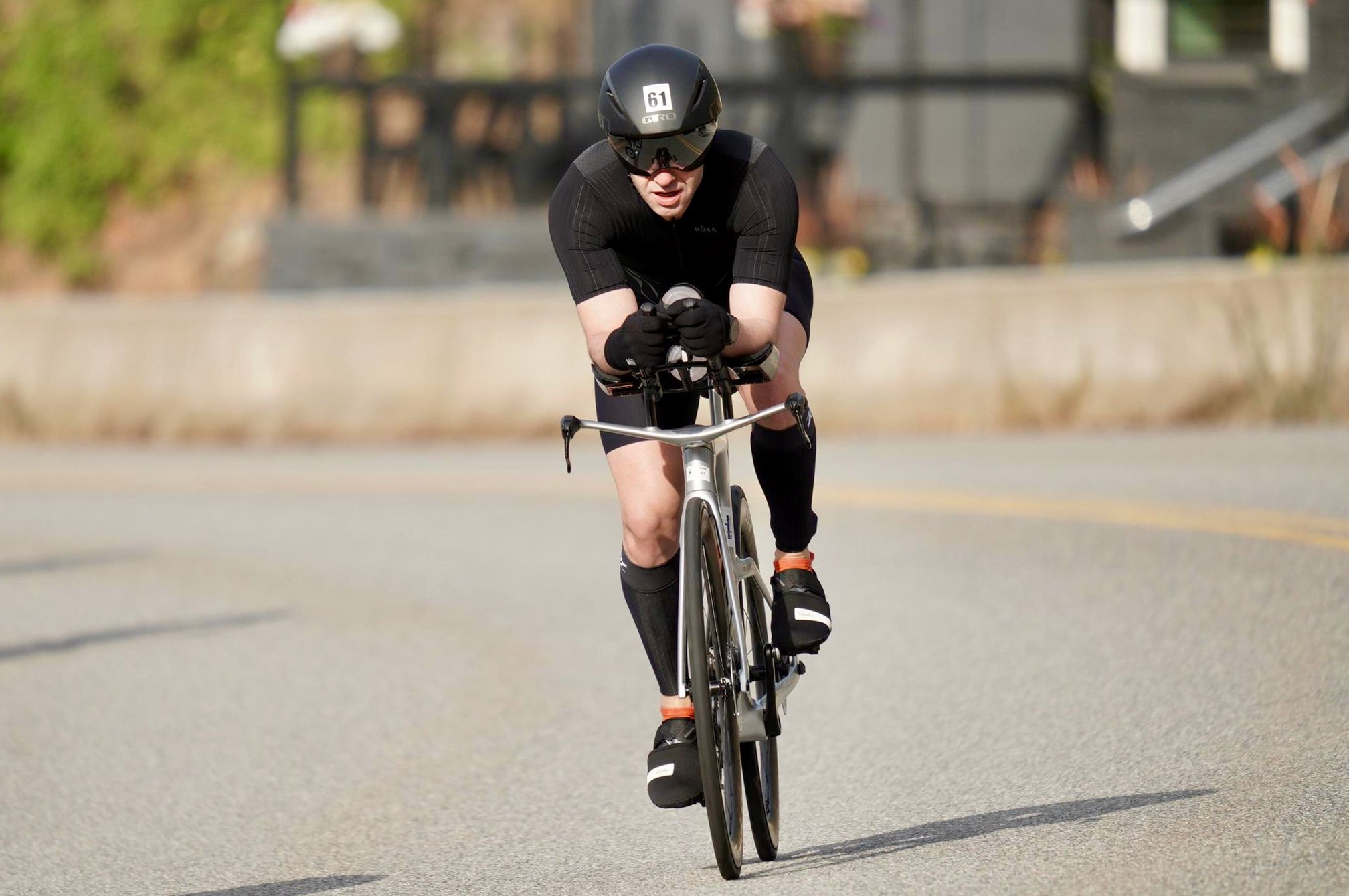 A frontal view of Guillermo Esteves in black gear and helmet with race number 61, riding a silver Trek Speed Concept triathlon bike in aero position on a curving paved road.