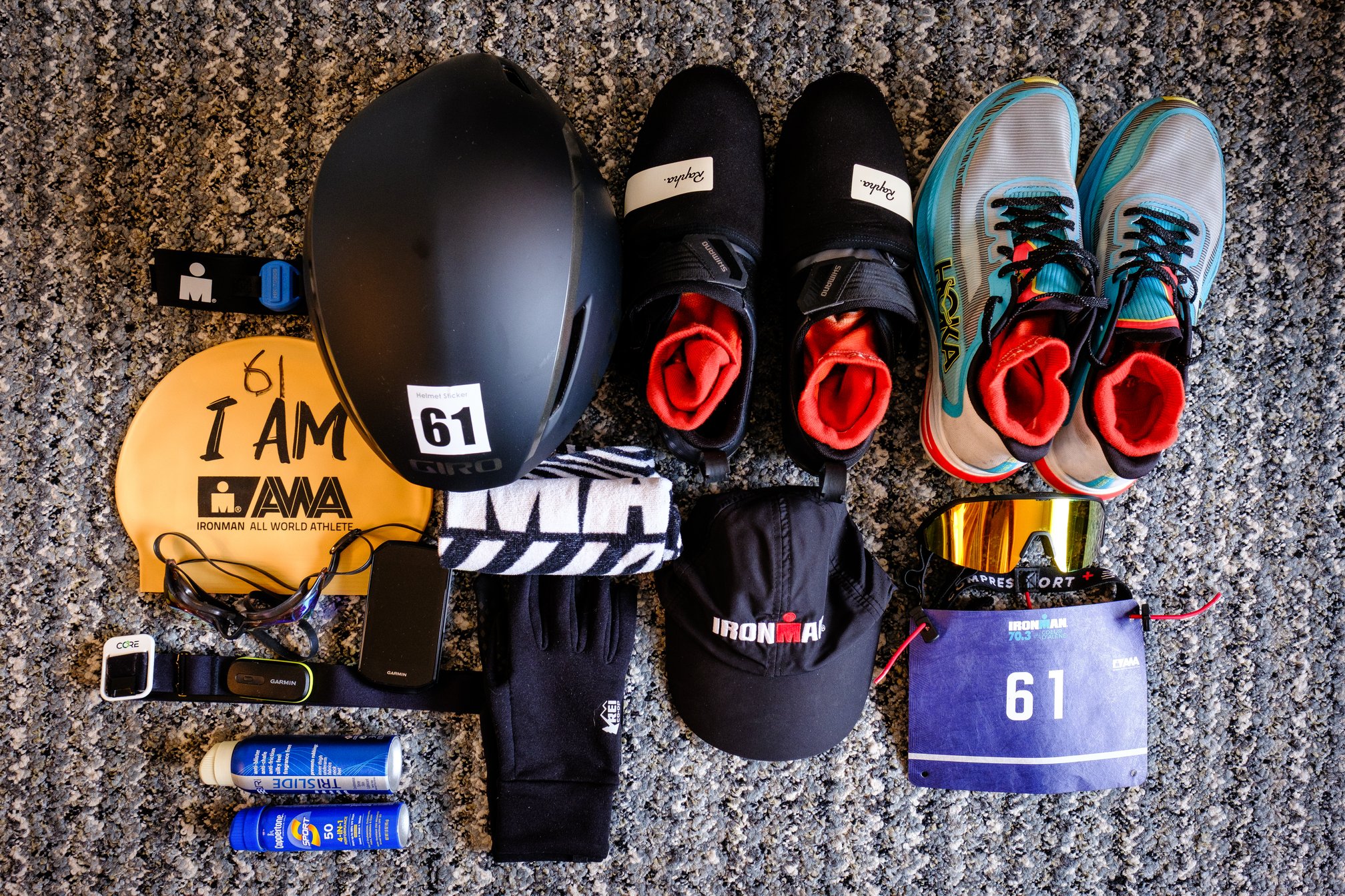 Triathlon gear laid out on a carpet, including an ankle timing chip, a swim cap with "I am AWA" printed on it and the number 61 written with a marker, a pair of swim goggles, a Garmin HRM with a Core body temperature sensor attached to the strap, a Giro Aerohead bike helmet with a 61 sticker, cycling shoes with toe covers on and running shoes (both with red socks rolled up inside), a towel, a Garmin 1050 bike computer, REI gloves, sunscreen, a tube of anti-chafe TriSlide spray, a black Ironman hat, a pair of golden sports sunglasses, and a race bib number 61 on a race belt.