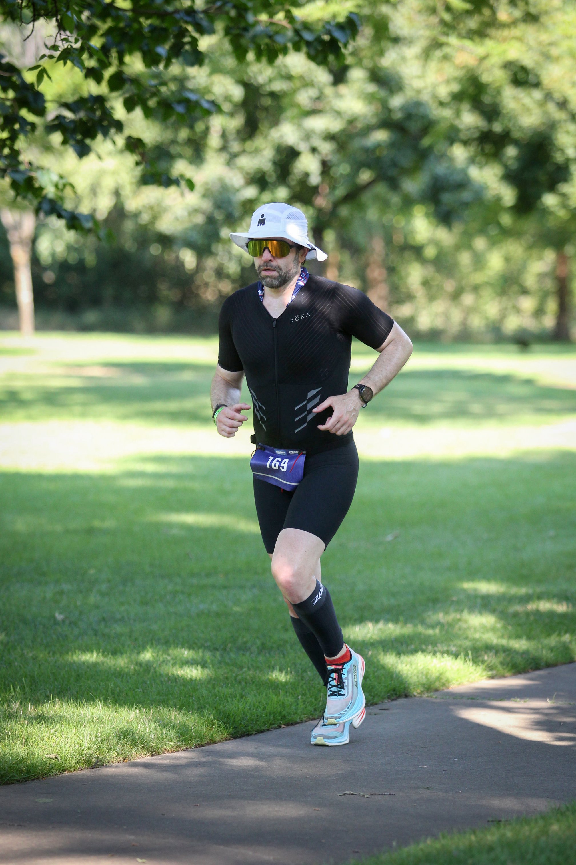 Guillermo Esteves, wearing a white boonie hat, golden sunglasses, and black trisuit with race bib number 169, running on a paved path through a park with green grass and trees in the background.