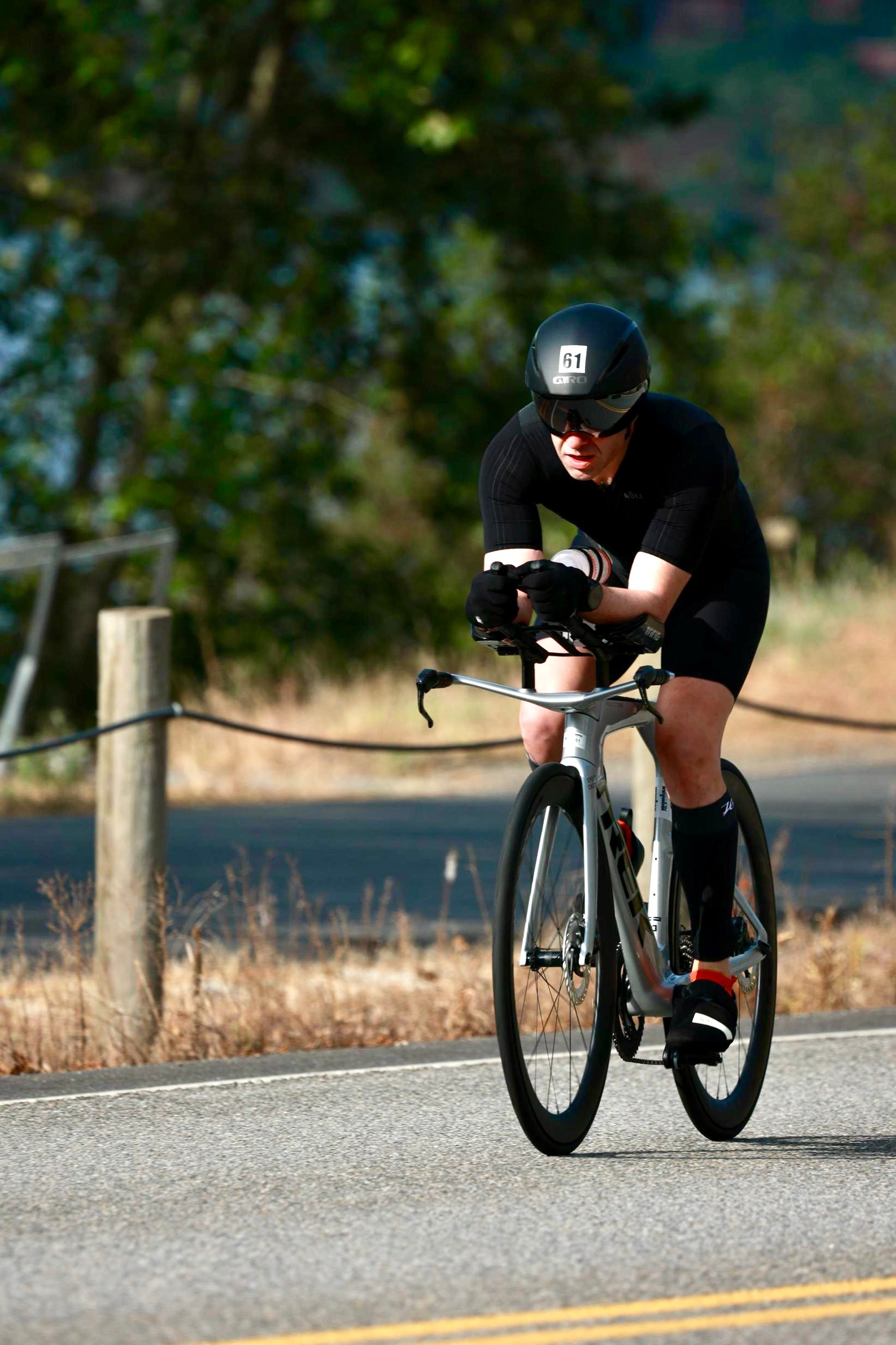 Guillermo Esteves, wearing black triathlon gear and race bib number 61, riding a silver Trek Speed Concept bike in an aerodynamic position on a paved road.