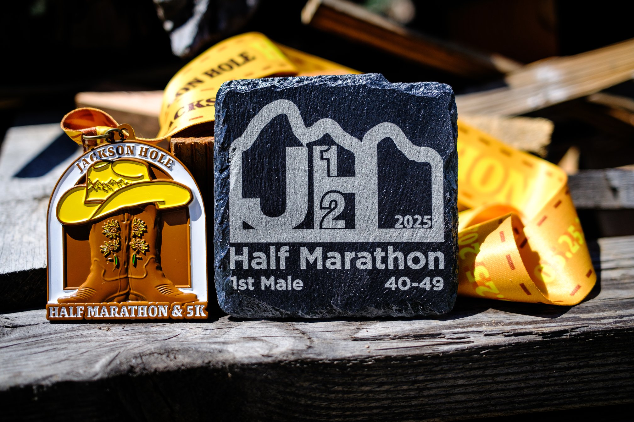 Two awards set on a pile of wooden scraps. On the left, there is a medal with a yellow ribbon that reads "Jackson Hole Half Marathon & 5K," and features two cowboy boots with a cowboy hat on top. On the right, there is a square slate plaque that reads "JH 1/2 2025" within a stylized silhouette of the Teton Range. Underneath, it reads "Half Marathon 1st Male 40–49." The medal's yellow ribbon, printed with "Jackson Hole," is visible behind the awards.