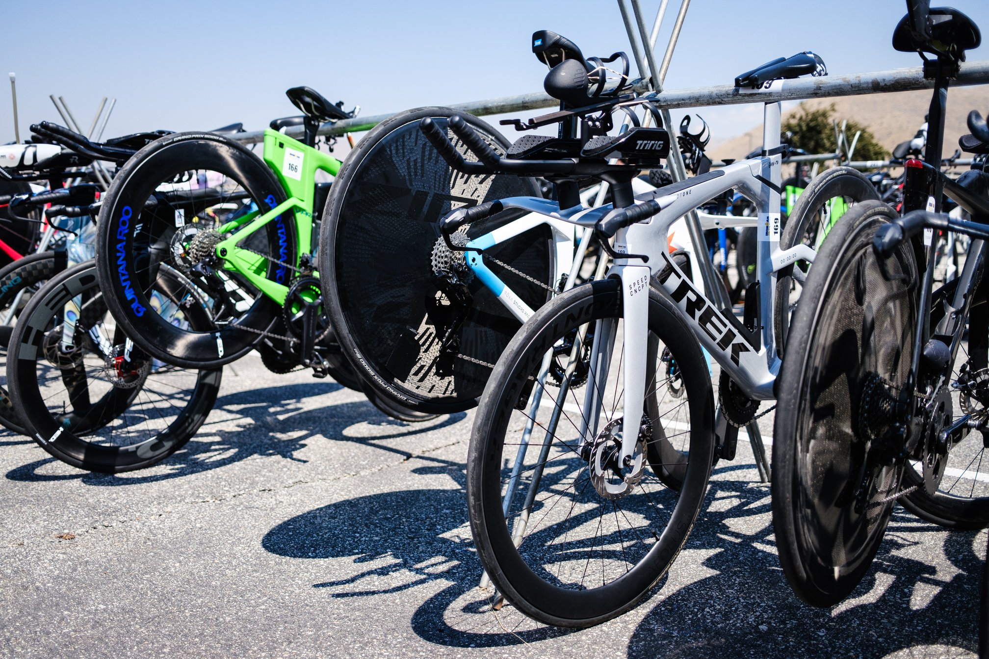 Multiple triathlon bicycles racked in a transition area in a parking lot, including a silver Trek Speed Concept SLR 7 in the foreground.