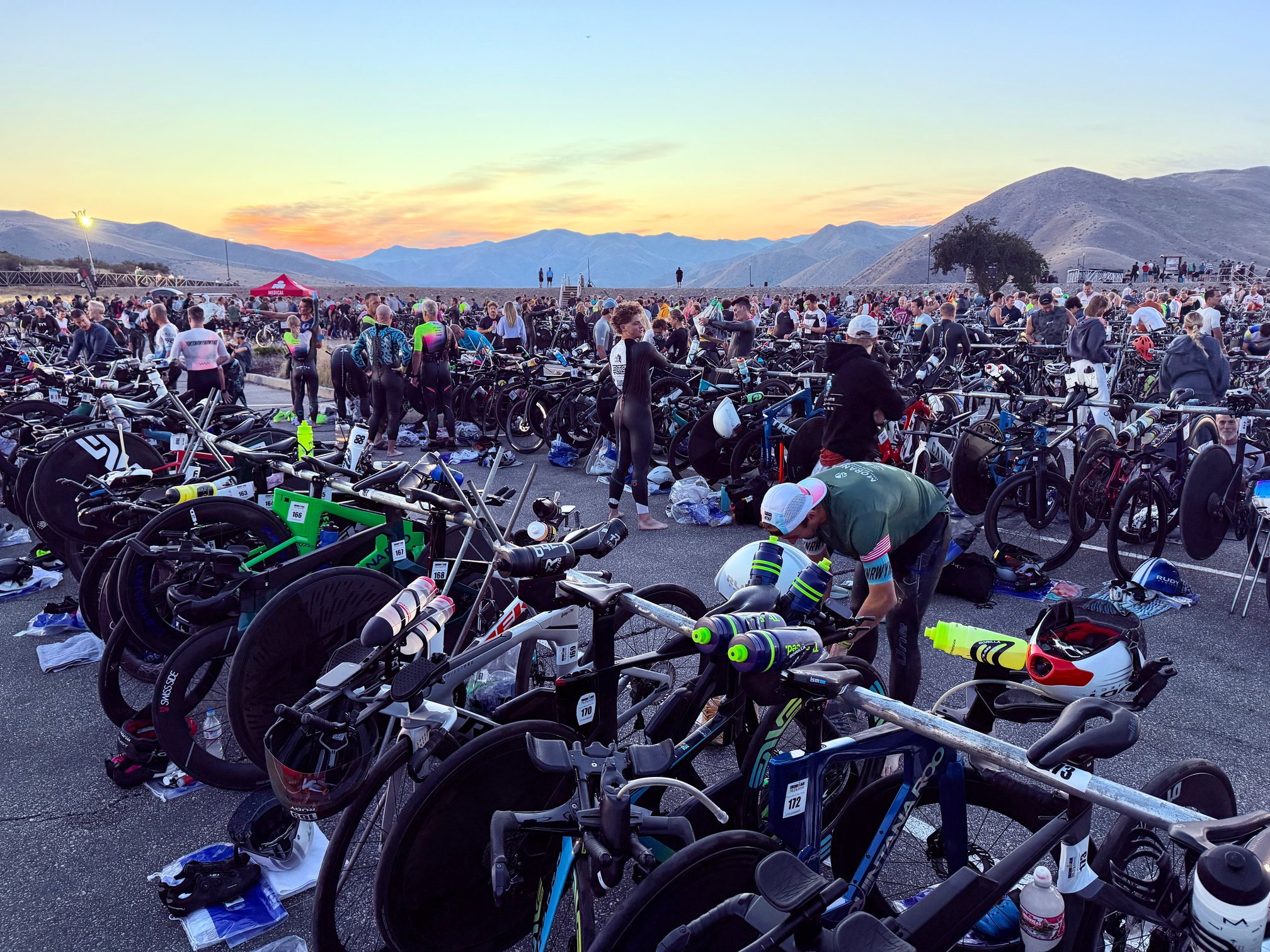 The T1 transition area of Ironman 70.3 Boise at sunrise, with hundreds of bikes racked in a paved parking lot and athletes setting up their gear and donning wetsuits.